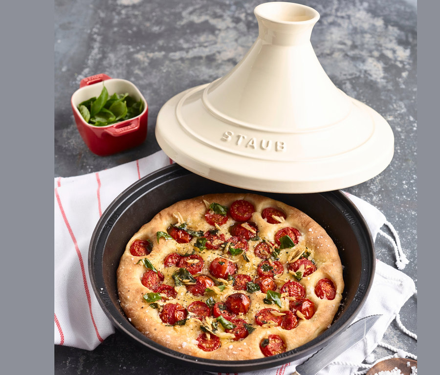 Focaccia mit Tomaten und Basilikum in einer schwarzen STAUB Gusseisen-Tajine, mit Deckel und einer roten Schale Basilikum.