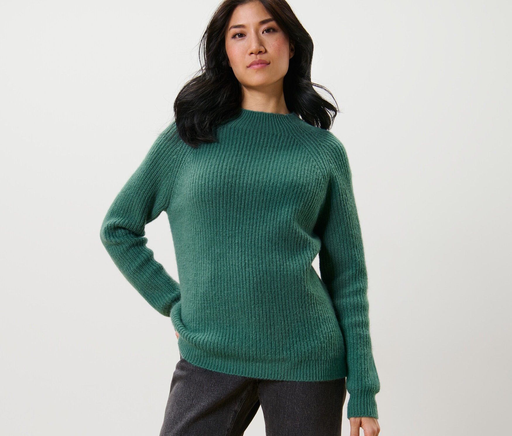 Frau posiert in grünem Grobstrickpullover mit Stehkragen und dunkler Hose.
