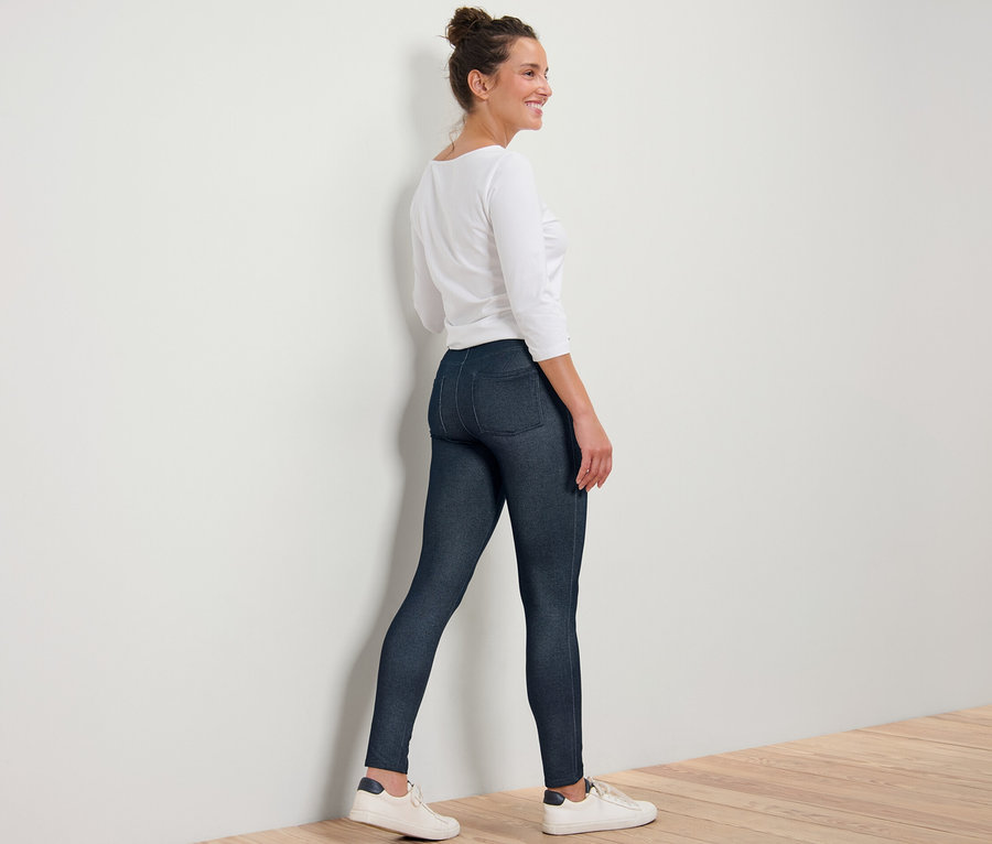 Eine Frau in Leggings in Jeansoptik, einem weißen Oberteil und Turnschuhen steht an einer Wand.