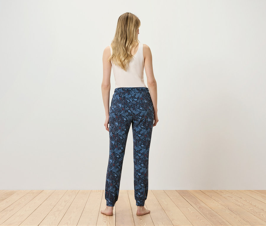 Frau steht mit dem Rücken zur Kamera und trägt ein weißes Top und eine blaue Hose mit Muster.