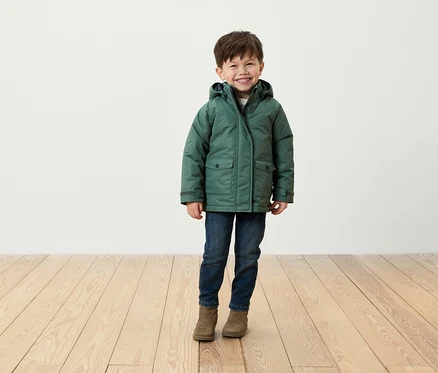 Ein lächelnder Junge steht auf einem Holzboden und trägt eine grüne Jacke, Jeans und braune Stiefel.