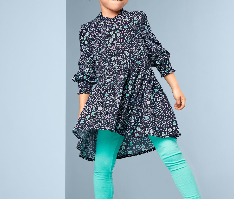 Lächelndes Mädchen posiert in einem Webkleid mit Blumenmuster, türkisfarbenen Leggings und weißen Turnschuhen.