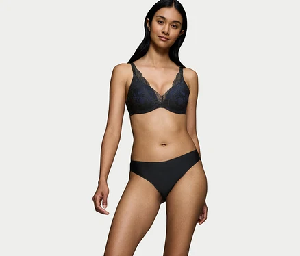 Frau mit dunklem Haar trägt einen schwarzen Triumph Body Makeup Illusion Lace WP BH mit blauer Fütterung und schwarze Slips vor weißem Hintergrund.