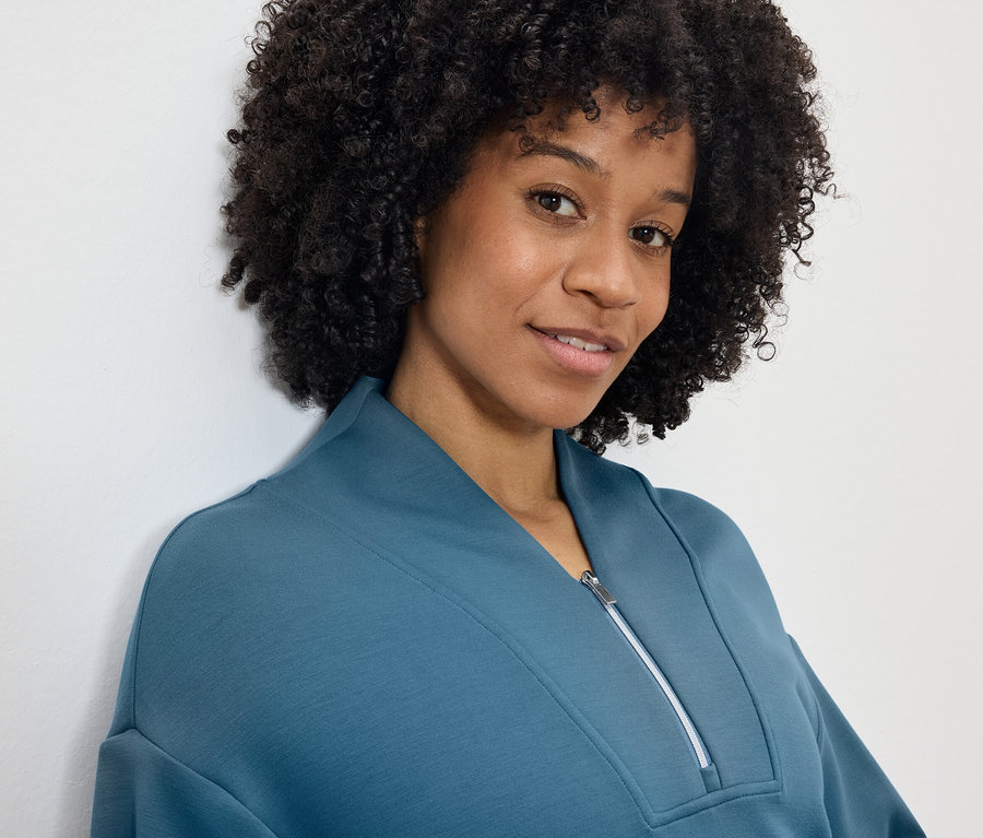 Porträt einer lächelnden Frau mit Afro-Frisur in einem blaugrünen Sweatshirt mit Reißverschluss.