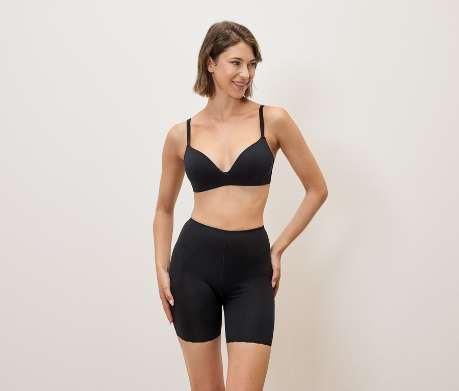 Frau mit schwarzem BH und schwarzen Shorts posiert vor einem neutralen Hintergrund.