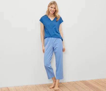 Blonde Frau steht und trägt ein Pyjama-Set, bestehend aus einem blauen T-Shirt und blau-weiß gestreiften Hosen.