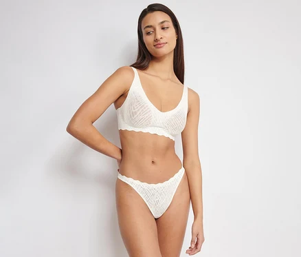 Eine Frau mit langen dunklen Haaren trägt einen weißen sloggi ZERO Feel Bliss Soft Bra und passende Slips. Der BH und die Slips haben eine dezente Mustertextur und gewellte Kanten. Sie steht mit einer Hand an der Hüfte und blickt zur Seite.