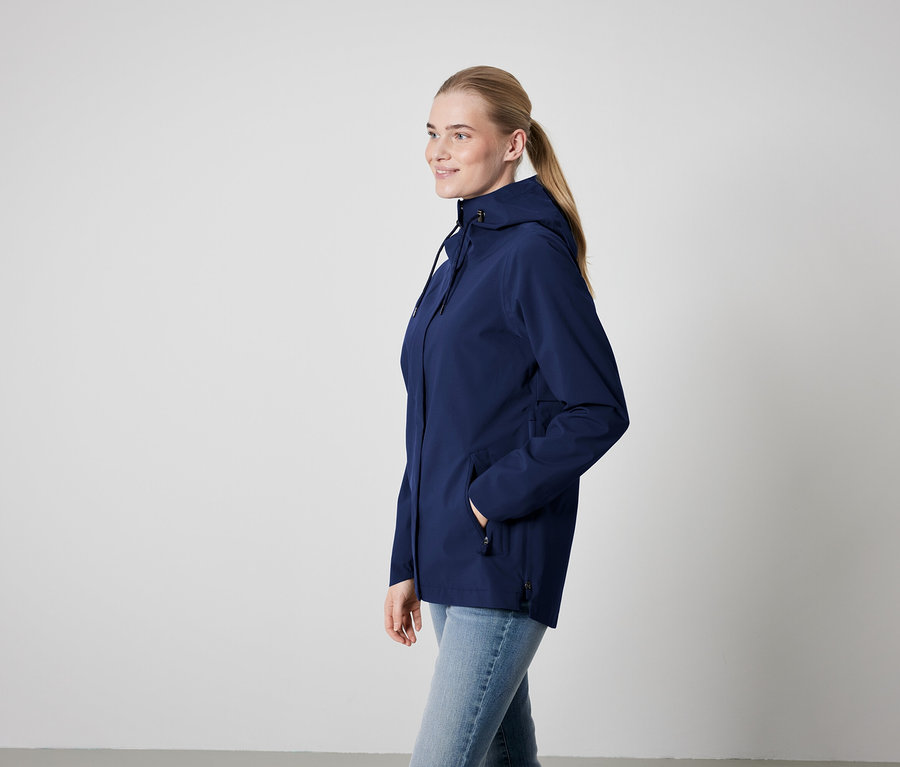 Frau in blauer Kapuzenjacke und Jeans steht seitlich.