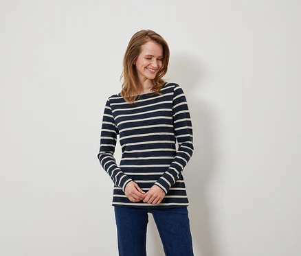 Eine Frau mit braunen Haaren und blauen Augen trägt ein blau-weiß gestreiftes Langarmshirt und blaue Jeans.