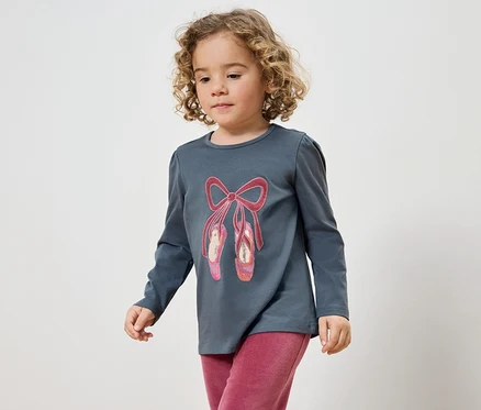 Mädchen mit lockigem Haar trägt ein graues T-Shirt mit einem Ballett-Schuh-Aufdruck und eine pinke Hose.