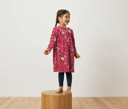 Ein lächelndes Mädchen steht auf einem Holzpodest und trägt ein rotes Langarmkleid und dunkelblaue Leggings.
