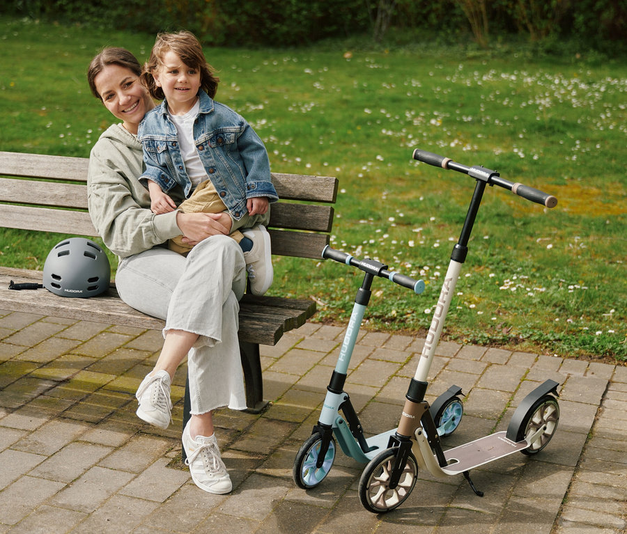 Eine Frau sitzt mit einem Kind auf einer Bank im Park. Daneben stehen zwei HUDORA BigWheel 180 Advanced Junior Roller und ein grauer Helm.