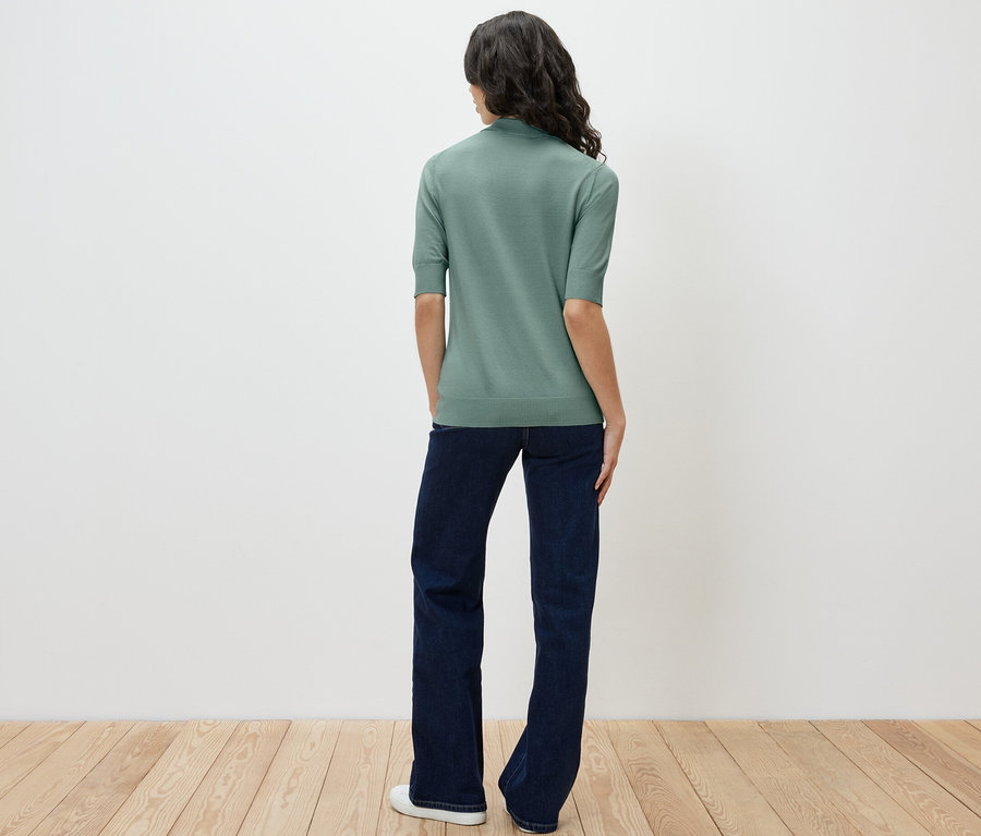 Eine Frau steht mit dem Rücken zur Kamera und trägt ein hellgrünes Feinstrick-Polo und Jeans.