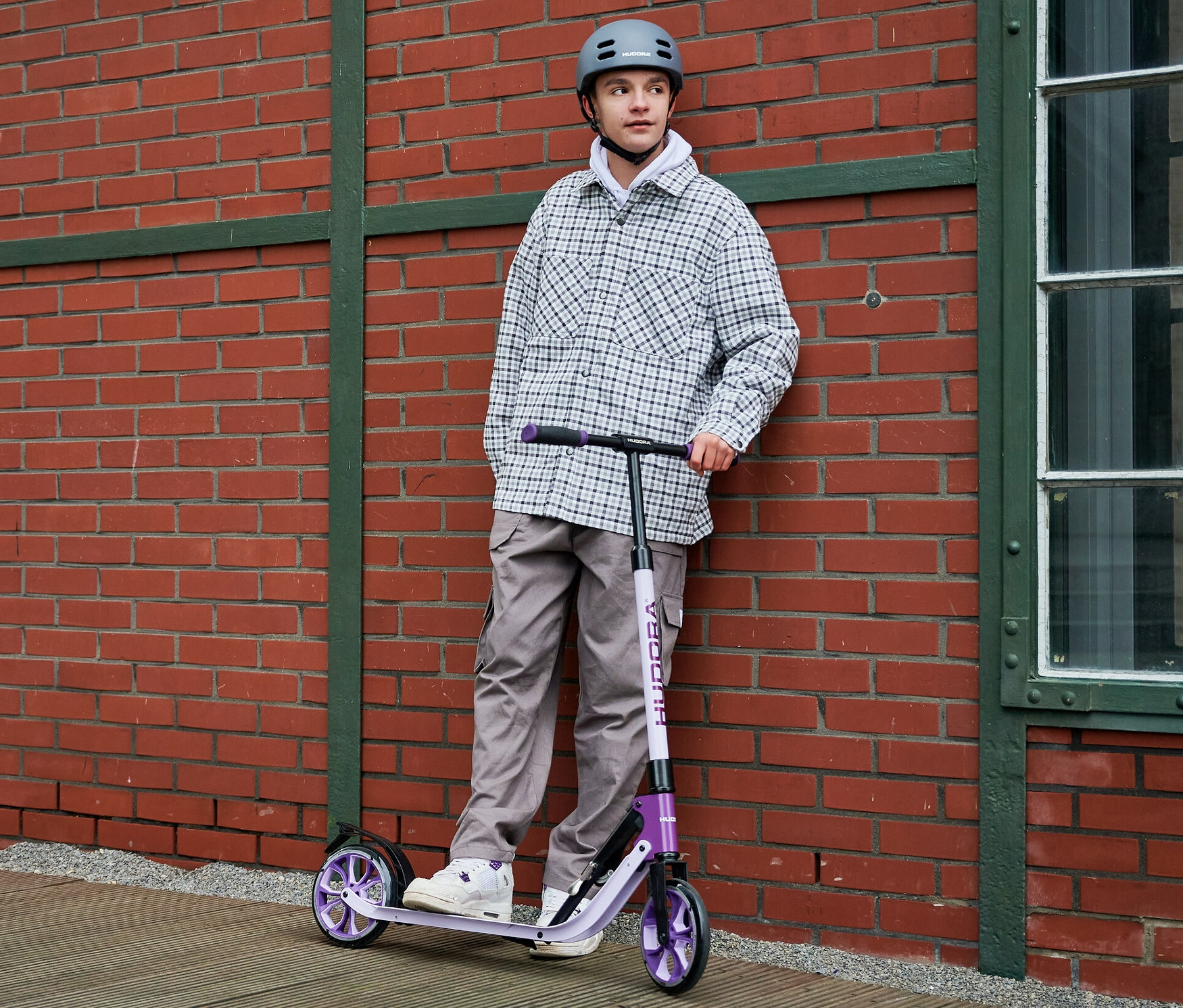 Eine Person mit grauem Helm steht an einer Ziegelwand mit einem lavendelfarbenen HUDORA BigWheel® 205 advanced Scooter.