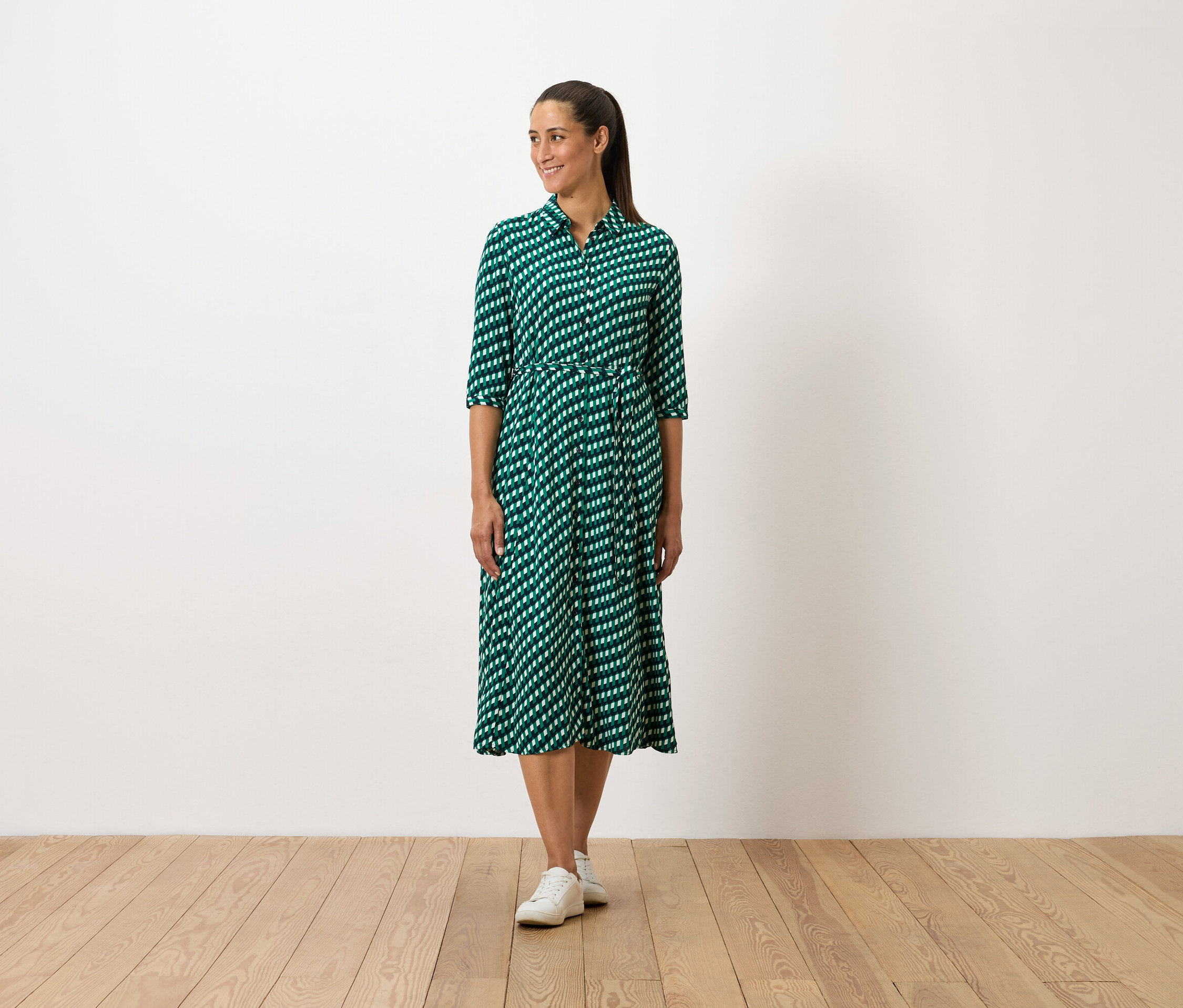 Eine Frau steht und trägt ein grünes gemustertes Kleid und weiße Turnschuhe.