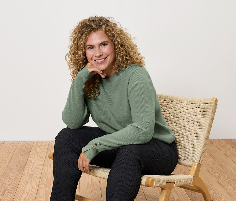 Frau sitzt auf einem Stuhl mit grünem Pullover und schwarzen Hosen.