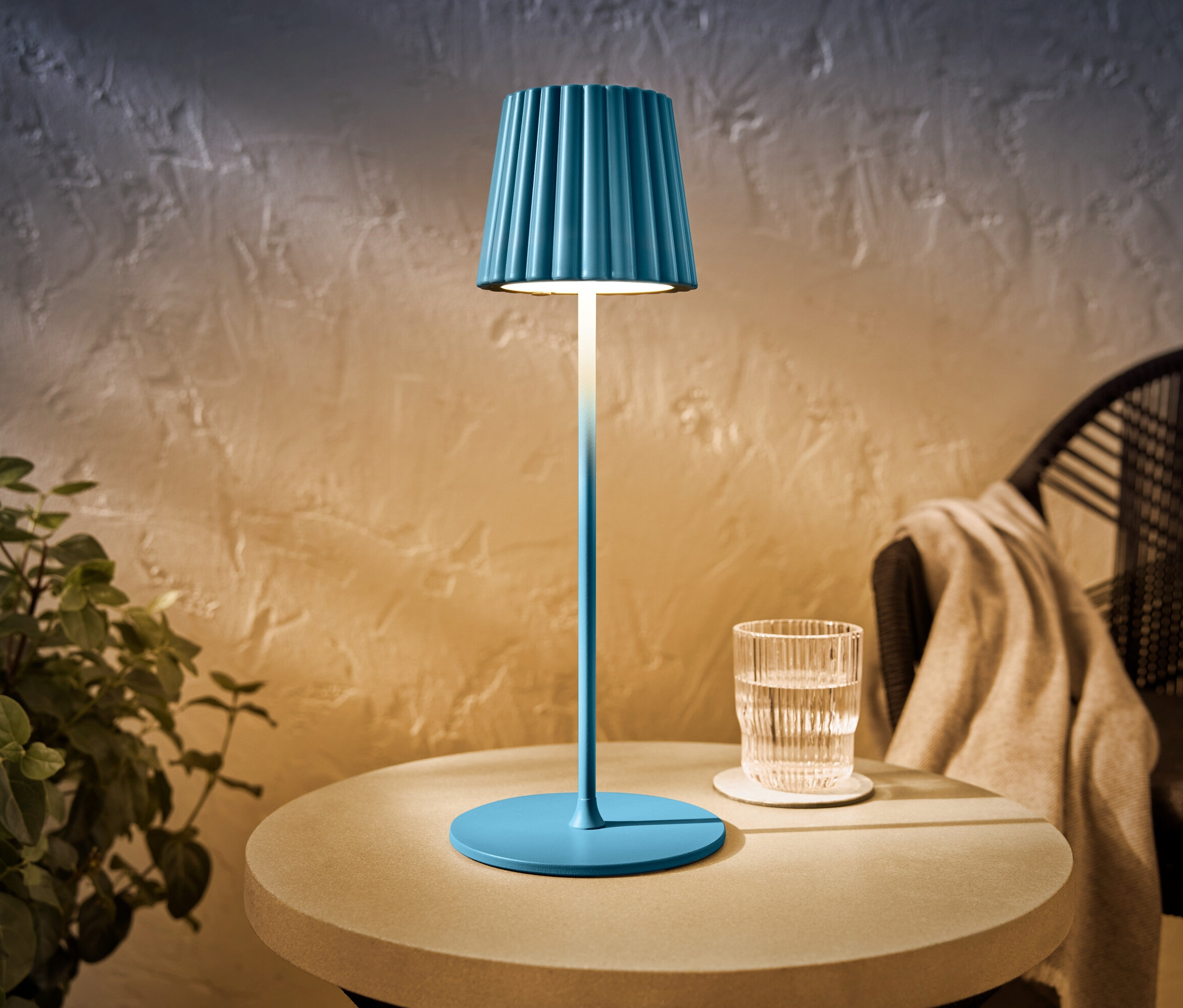 Eine blaue LED-Akku-Outdoor-Tischleuchte steht auf einem Tisch neben einem Glas Wasser.