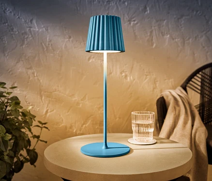 Eine blaue LED-Akku-Outdoor-Tischleuchte steht auf einem Tisch neben einem Glas Wasser.