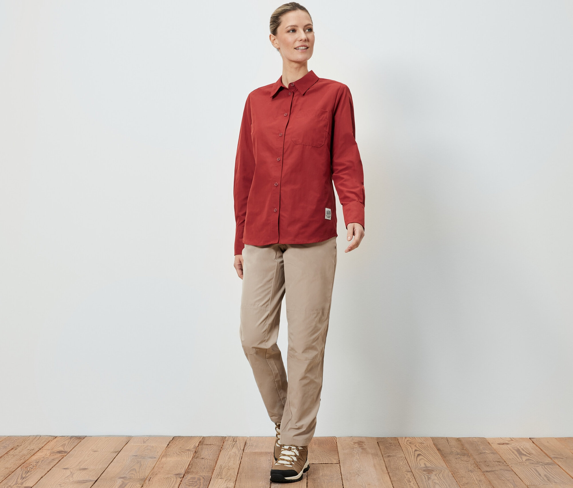 Eine Frau in einer Outdoorbluse mit Moskito-Schutz, beigen Hosen und Wanderschuhen steht auf einem Holzboden vor einer weißen Wand.