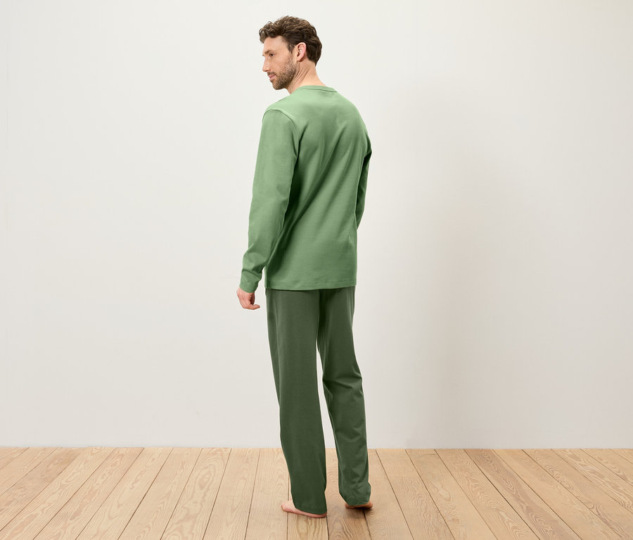 Ein Mann steht mit dem Rücken zum Betrachter und trägt einen grünen Schlafanzug auf einem Holzboden.