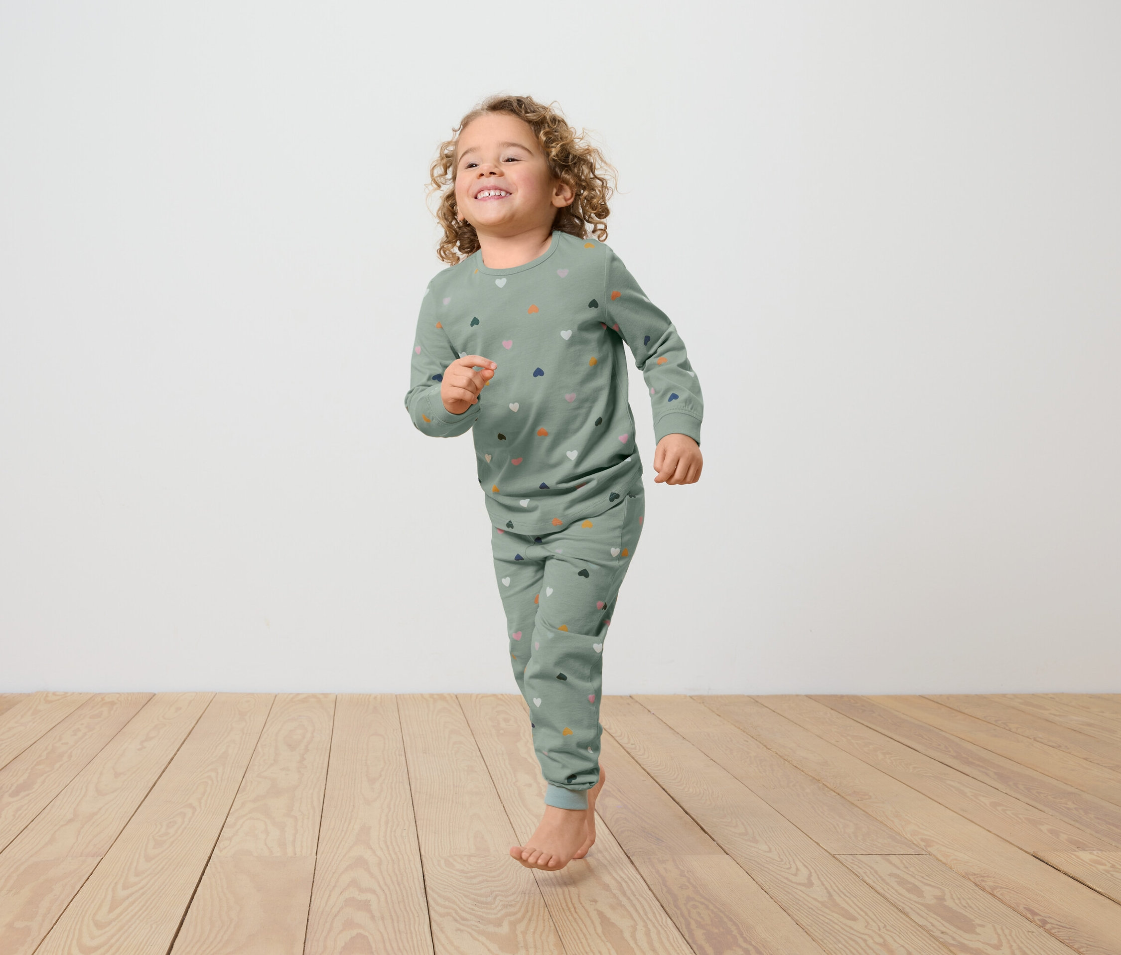 Mädchen mit lockigem Haar, bekleidet mit einem grünen Pyjama mit Herzmuster, rennt auf einem Holzboden.