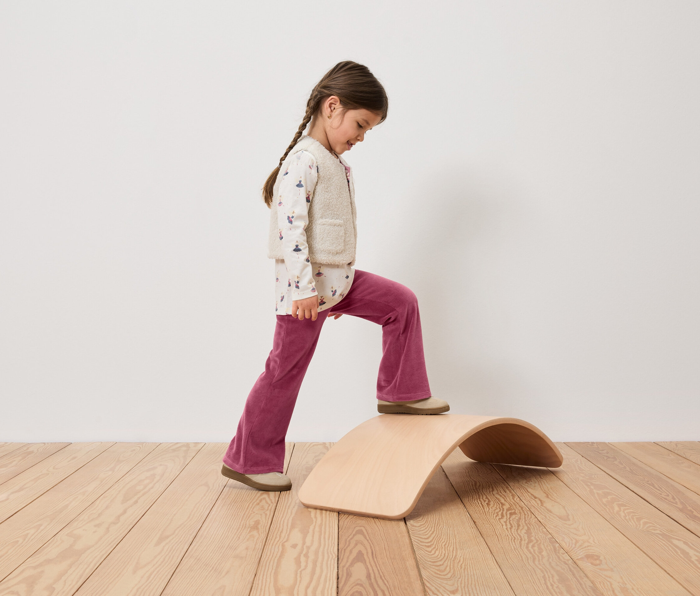 Mädchen mit Zopf steigt auf ein hölzernes Balance Board, gekleidet in rosafarbene Cordhose, bedrucktes Oberteil und Weste.