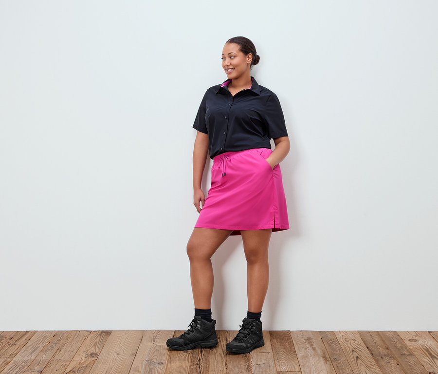 Eine Frau steht vor einer weißen Wand, sie trägt ein schwarzes Hemd, einen pinken Rock und schwarze Schuhe.