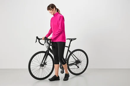 Frau mit pinkfarbener Jacke, schwarzen Radlerhosen und schwarzen Schuhen, die neben einem schwarzen Fahrrad steht.