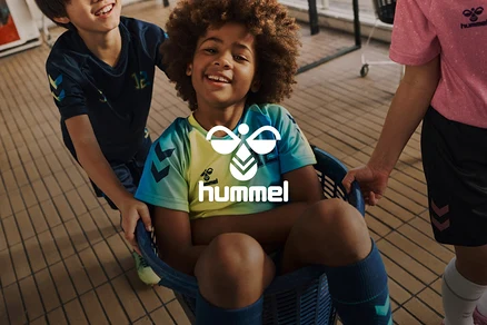 Drei Kinder, davon eines in einem Korb sitzend, tragen Hummel-Sportbekleidung und lächeln.