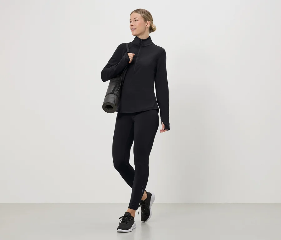 Frau in schwarzem Sportoberteil, Leggings und Turnschuhen mit Yogamatte über der Schulter.