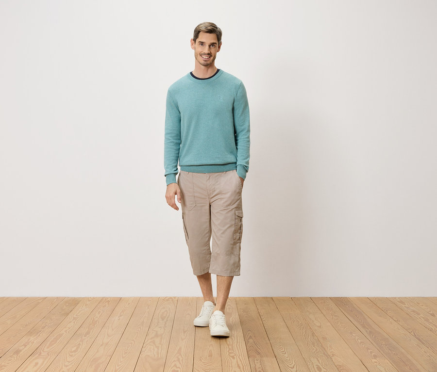 Ein Mann steht mit einer Hand in der Tasche vor einer weißen Wand. Er trägt einen hellblauen Pullover, beigefarbene Dreiviertel-Cargo-Hosen und weiße Schuhe.