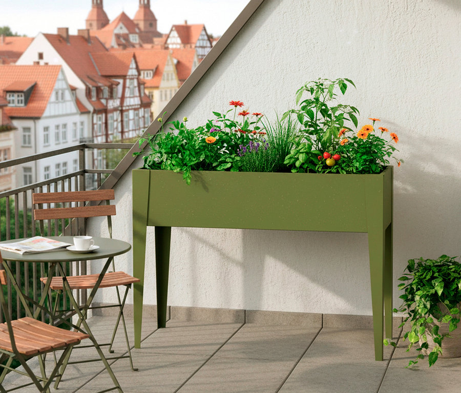 Auf einem Balkon steht ein grüner Pflanzkasten mit Blumen und Pflanzen, daneben ein Gartentisch mit zwei Stühlen.