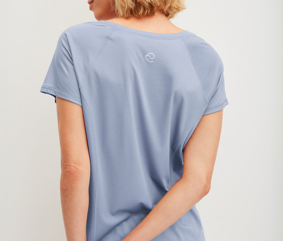 Frau mit kurzem blonden Haar, von hinten gesehen, trägt ein hellblaues T-Shirt mit kurzen Ärmeln und eine passende kurze Hose, die Hände hinter dem Rücken verschränkt.