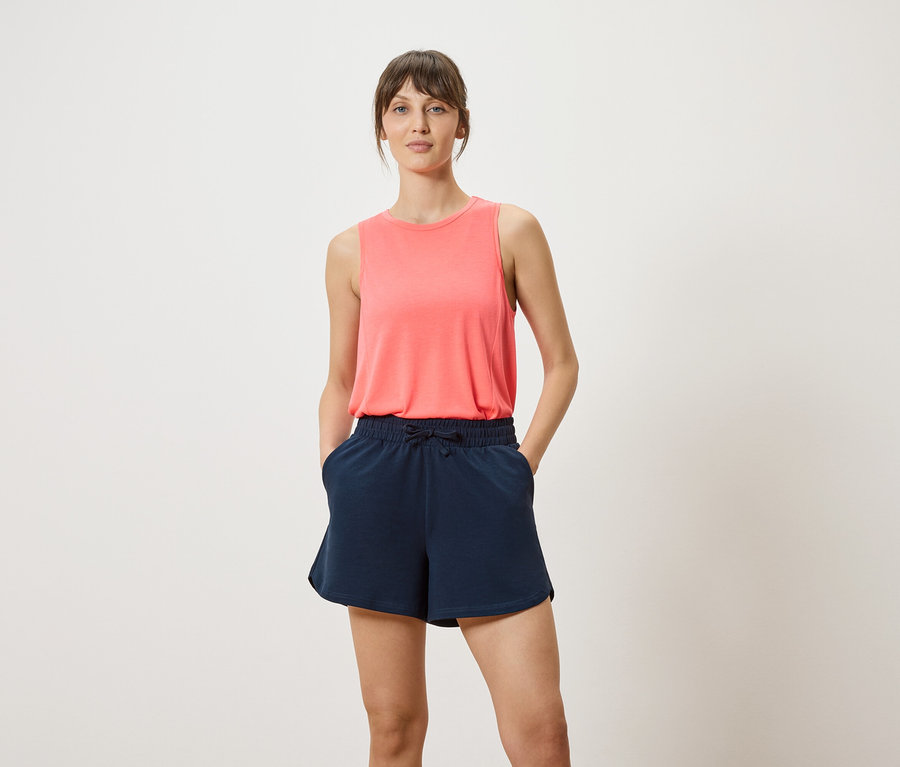 Frau mit korallfarbenem Tanktop und marineblauen Shorts mit ihren Händen in den Taschen.