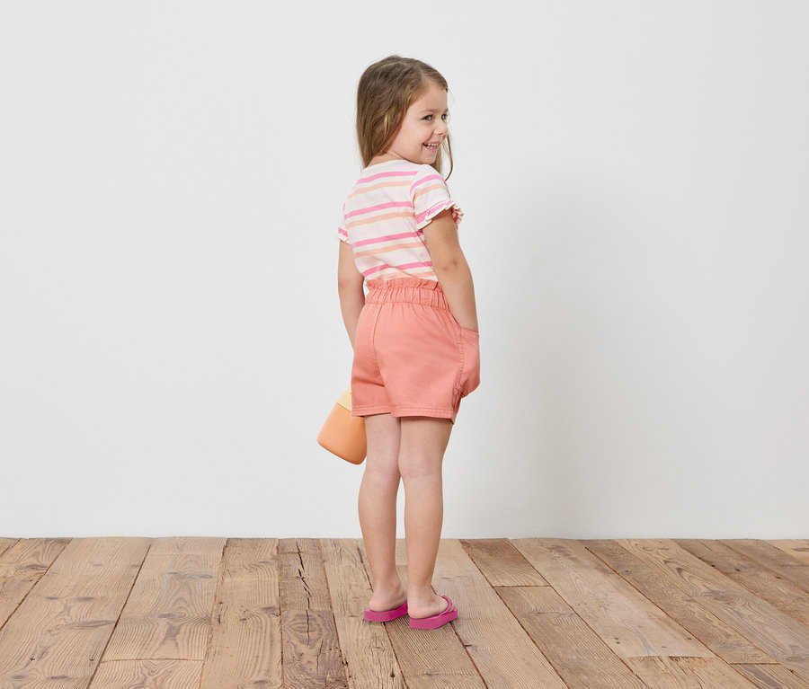 Mädchen steht mit dem Rücken zur Kamera, trägt Shorts, T-Shirt und Sandalen, hält einen Eimer in der Hand.