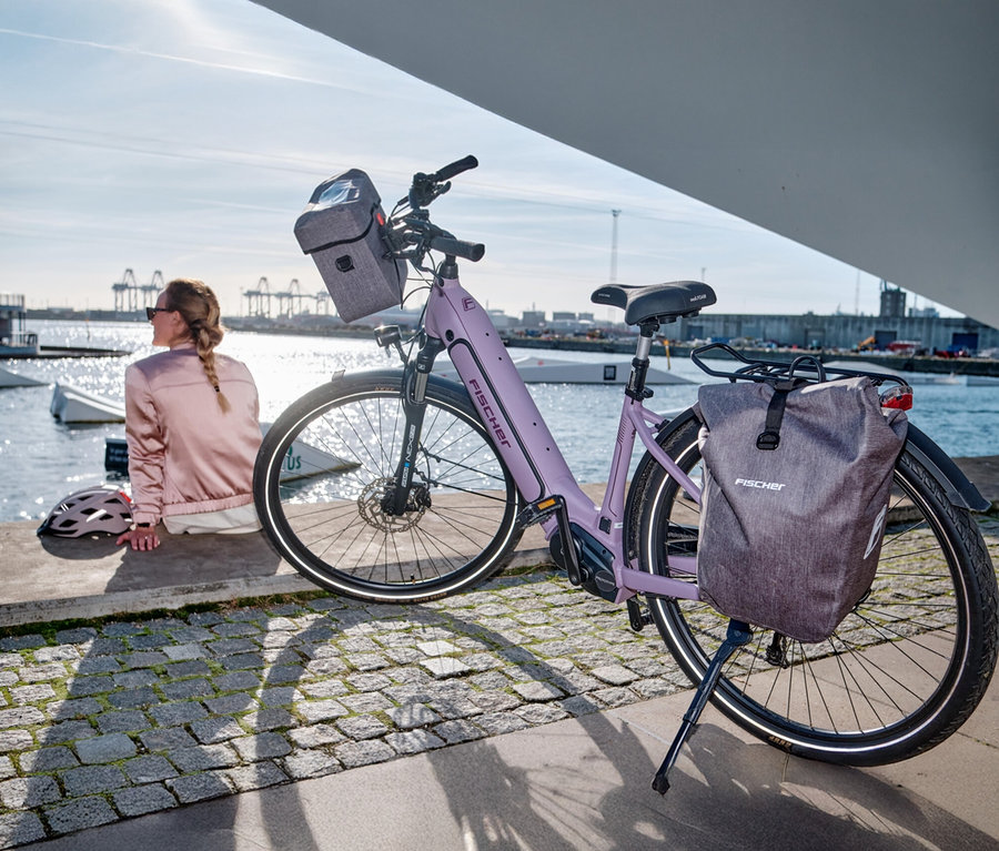 Fahrrad mit Fischer Gepäckträgertasche PLUS Cita steht am Kai, Frau sitzt daneben.