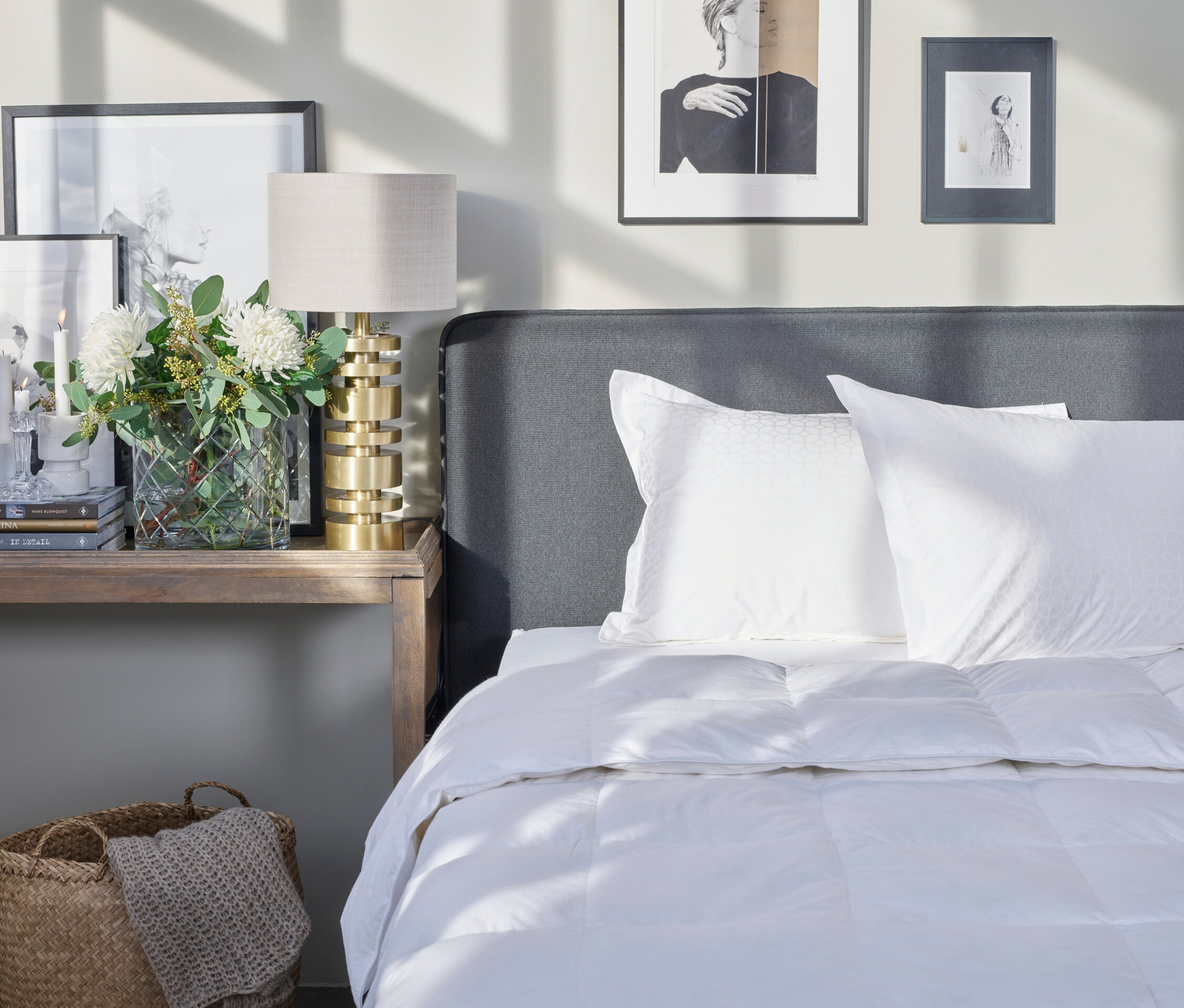 Schlafzimmer mit weißer Bettdecke, Kissen und grauem Kopfteil. Auf dem Nachttisch stehen eine Lampe, Blumen und gerahmte Bilder.