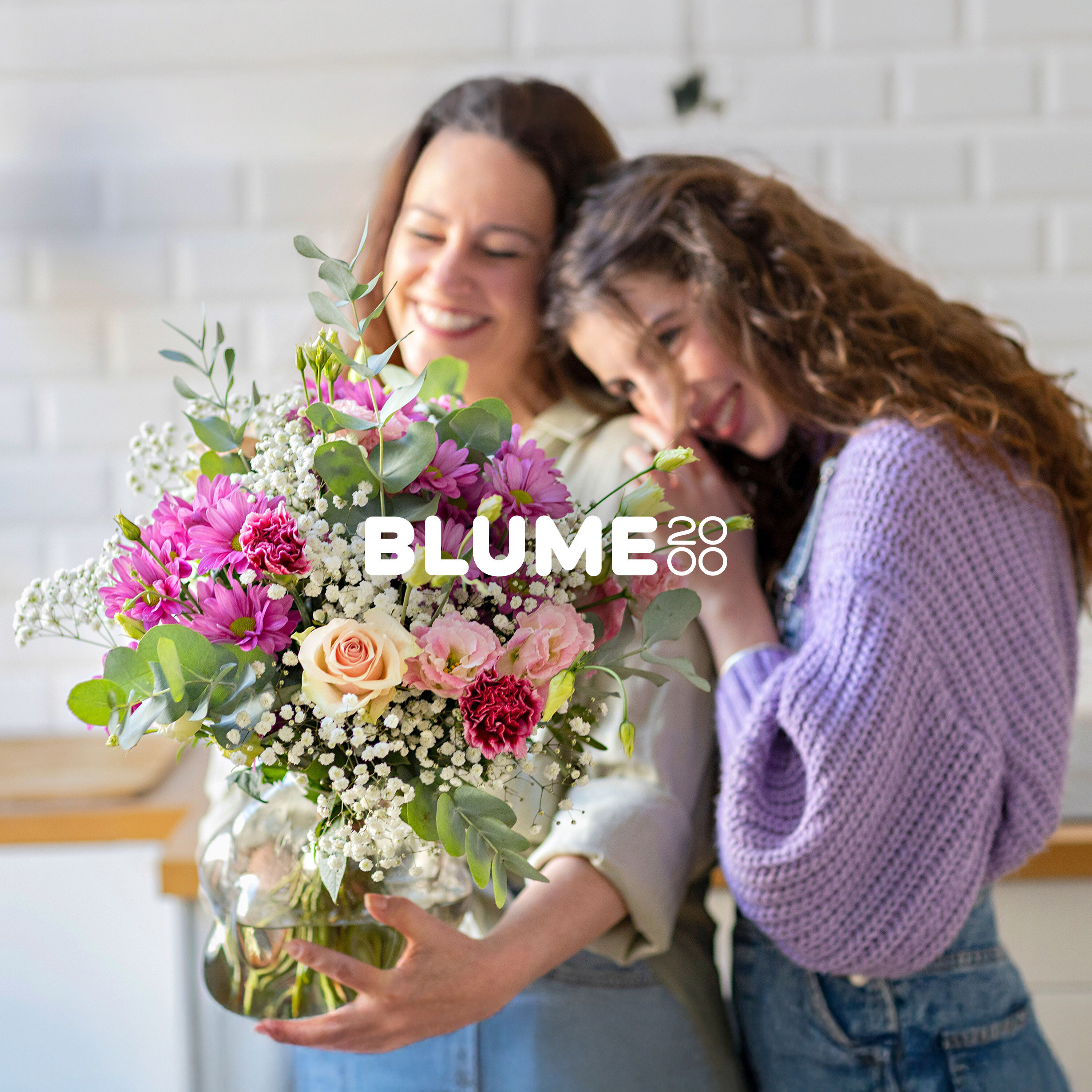Zwei lächelnde Frauen, eine hält einen lebhaften Strauß aus rosa, lila und cremefarbenen Blumen mit grünem Laub, während die andere ihren Kopf an ihre Schulter lehnt.