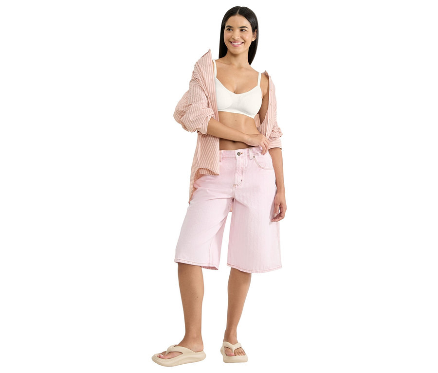 Lächelnde Frau trägt einen weißen sloggi ZERO Feel Pure Soft BH, ein offenes rosa gestreiftes Hemd, rosa Denim-Shorts und beige Flip-Flops.