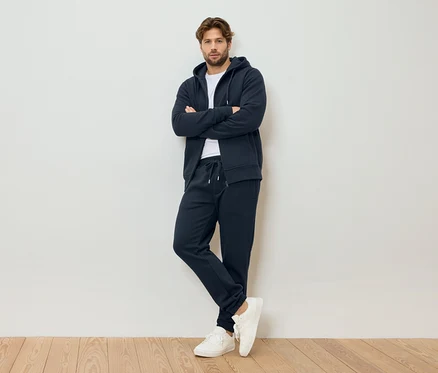 Mann mit verschränkten Armen, trägt marineblauen Kapuzenpullover mit Reißverschluss, marineblaue Jogginghose und weiße Turnschuhe, stehend vor einer weißen Wand.