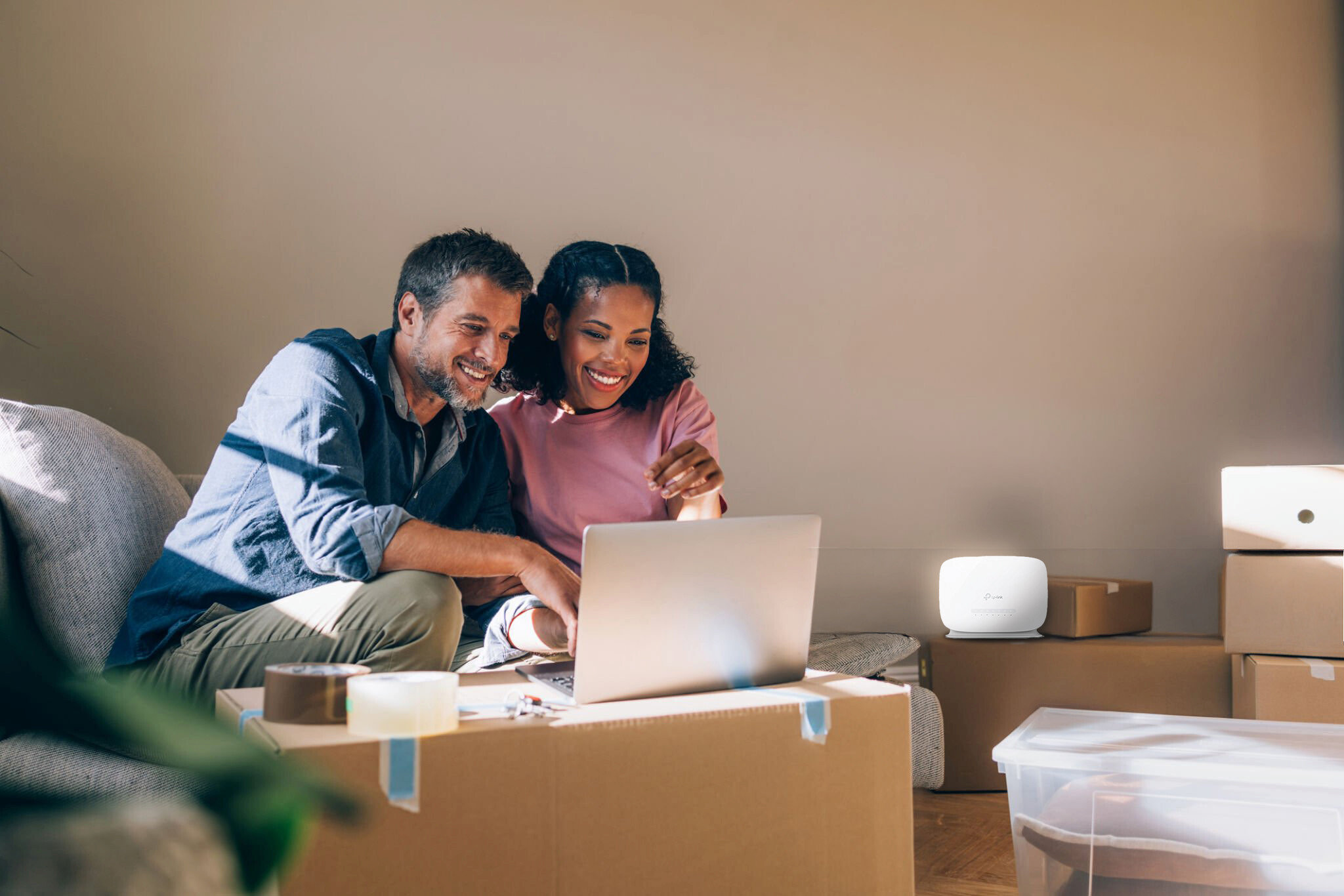 Ein Mann und eine Frau sitzen auf einem Sofa und schauen auf einen Laptop, umgeben von Umzugskartons.