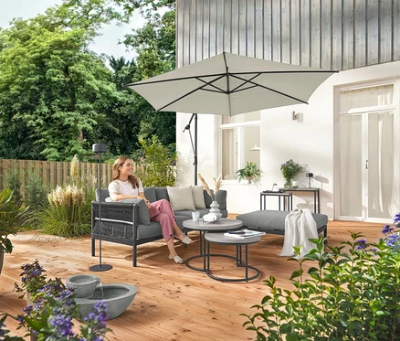 Eine Frau sitzt auf einem komfortablen Gartensofa mit separater Fußlehne. Eine graue Solar-Akku-Stehleuchte steht neben ihr. Im Vordergrund befindet sich ein Solar-Gartenbrunnen.