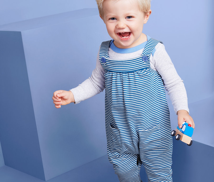 Lächelndes Kleinkind trägt eine Jersey-Latzhose und hält ein hölzernes Spielzeugauto.