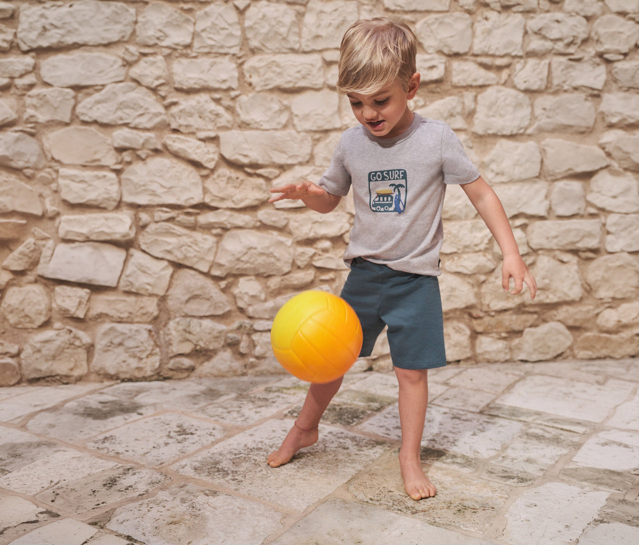Junge spielt barfuß mit gelbem Ball. Er trägt einen kurzen Kinder-Pyjama von SCHIESSER mit grauem T-Shirt und blauen Shorts.