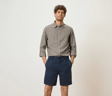 Mann mit grauem Hemd und blauen Shorts steht vor einem weißen Hintergrund.