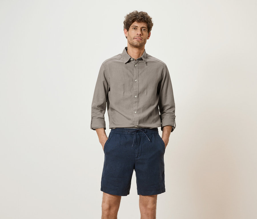 Mann mit grauem Hemd und blauen Shorts steht vor einem weißen Hintergrund.
