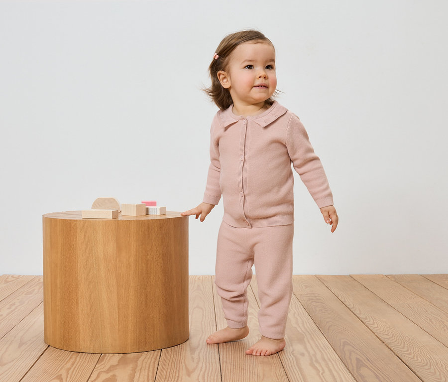 Kleinkind in rosa Strickjacke und Hose steht neben einem Holztisch mit Holzspielzeug auf einem Holzboden.