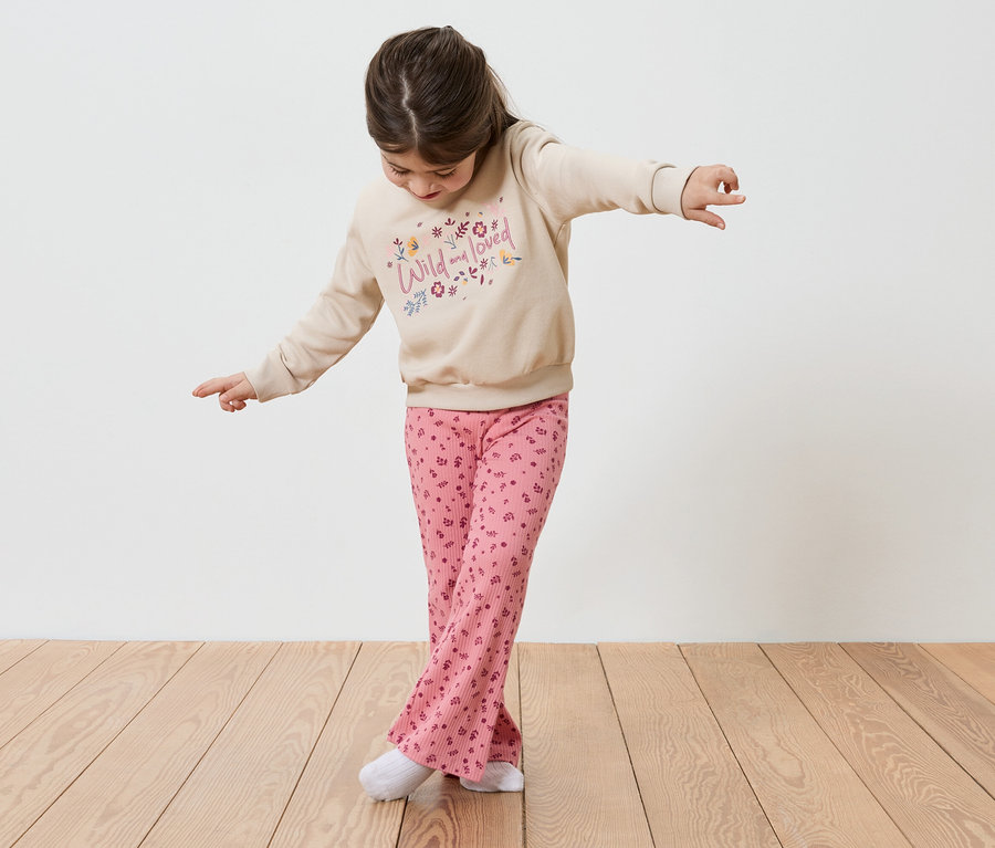 Ein Mädchen balanciert mit ausgestreckten Armen auf einem Holzboden. Sie trägt einen beigen Pullover mit der Aufschrift "Wild and loved" und rosafarbene Hosen.