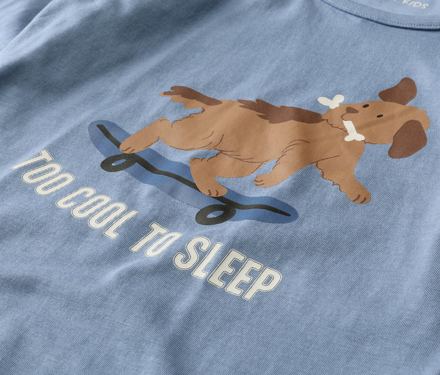 Hellblaues T-Shirt mit einem braunen Hund, der auf einem blauen Skateboard liegt und einen Knochen im Maul hält. Darunter steht der weiße Text "TOO COOL TO SLEEP".