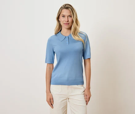 Blonde Frau trägt ein hellblaues Poloshirt und cremefarbene Hosen vor einem cremefarbenen Hintergrund.